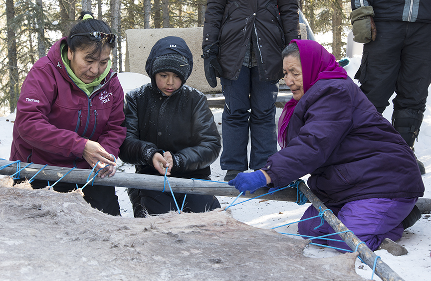 Therese Romie and Sophie Williah working with a student from Mezi Community School to attach a moosehide to a frame, Whati 2016. Photo: Fran Hurcomb