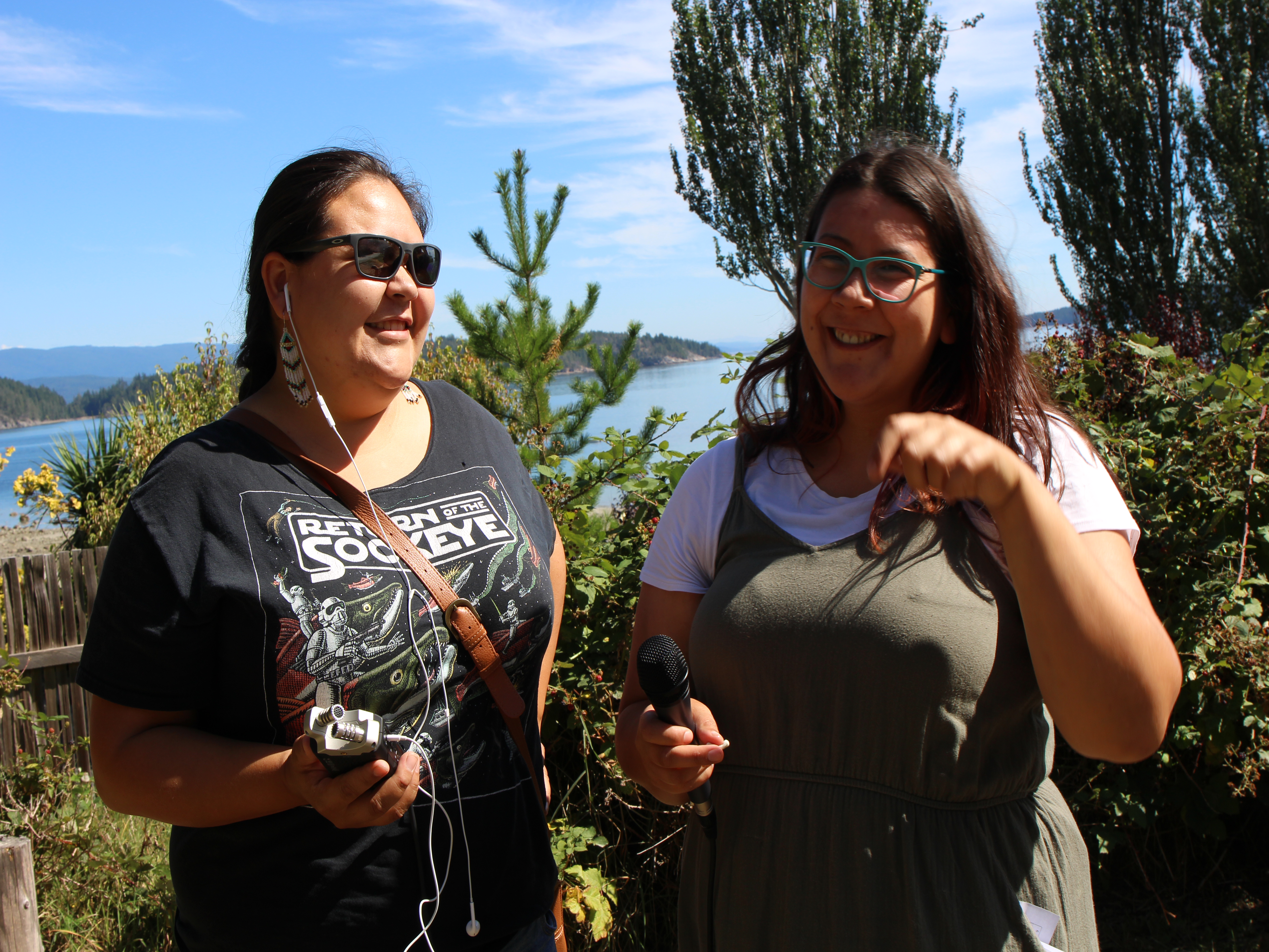 Two young women holding a recorder and microphone in front of a bush with a body of water in the background.