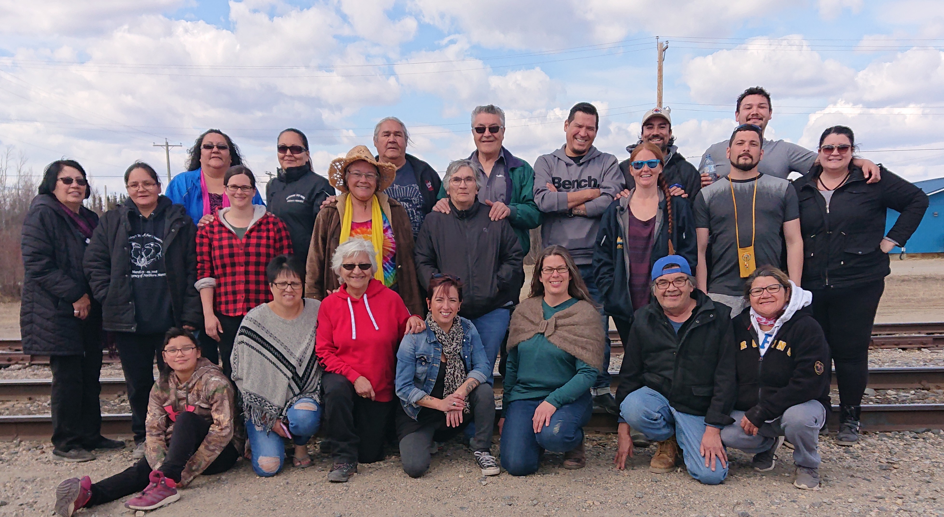 Group of NMFCCC members comprised of member of Indigenous communities in Northern Manitoba and others, standing outside in three rows with the front row kneeling.