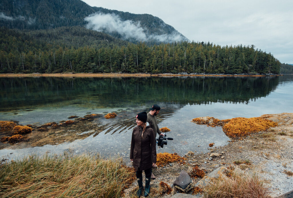 Emma Gilchrist, The Narwhal's Editor in Chief, stands at the edge of a body of water surrounded by forests in Bella Bella. She is wearing a black winter hat, a long rain jacket and black rain boots. Her blond hair is in a long braid and she is looking away from the camera. A cameraman stands behind her, looking in the opposite direction, video-recording the water.