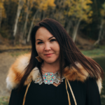 Lasänmą—Katie Johnson looks into the camera with a slight small on her face. She is wearing a large beaded necklace around her neck and a black coat that has fur around the hood. She is standing outside in front of birch trees that have turned golden.