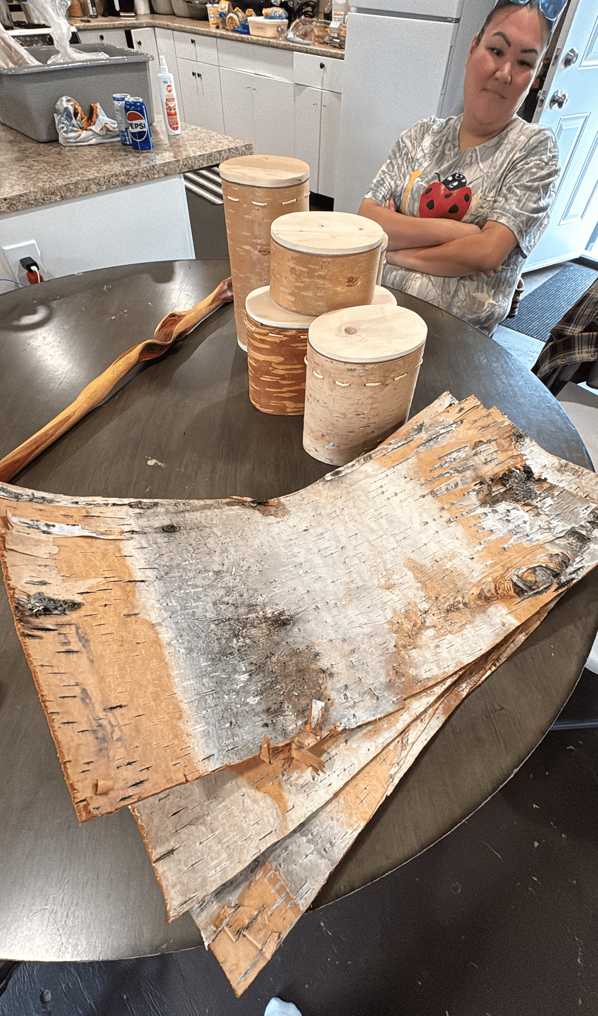 A woman sits at a table with her arms crossed. There are three sheets of birch bark on the table, as well as some birch bark baskets the group had made earlier.