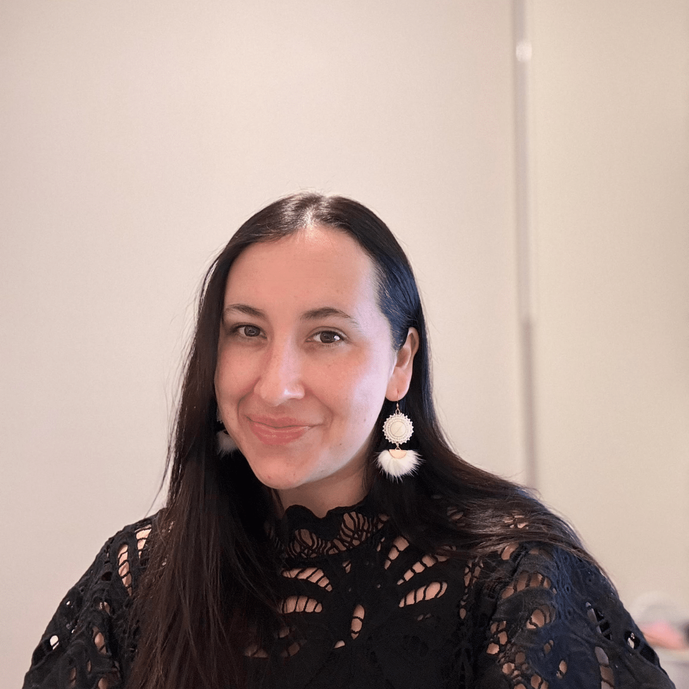 Emily Cabera seated indoors, wearing a black lace top and light‑colored statement earrings, with long dark hair, against a plain light background