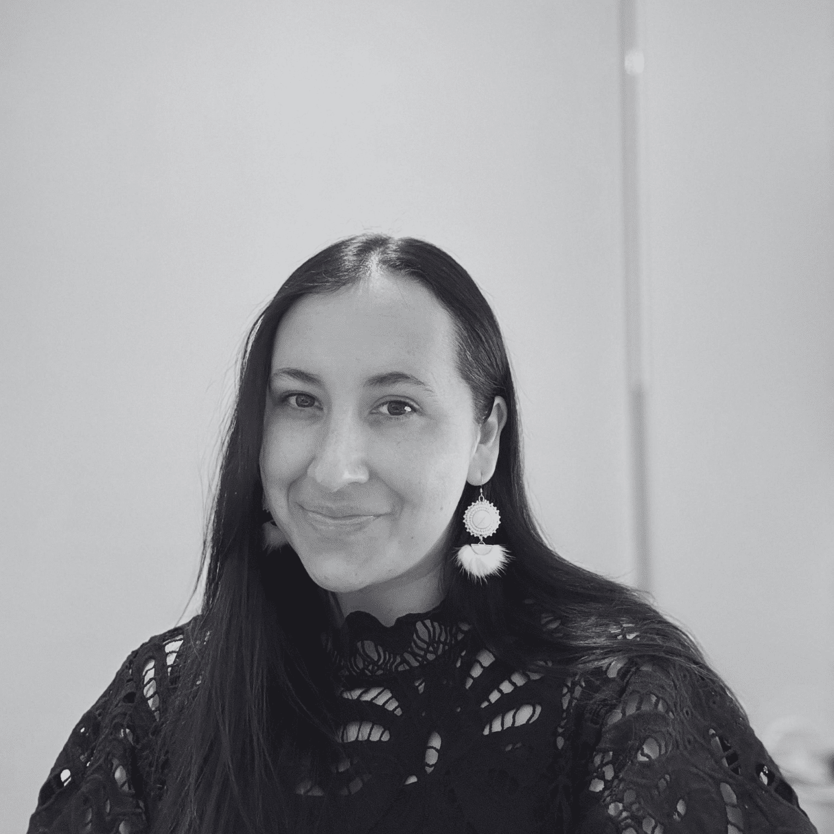 Emily Cabrera seated indoors, wearing a black lace top and light‑colored statement earrings, with long dark hair, against a plain light background