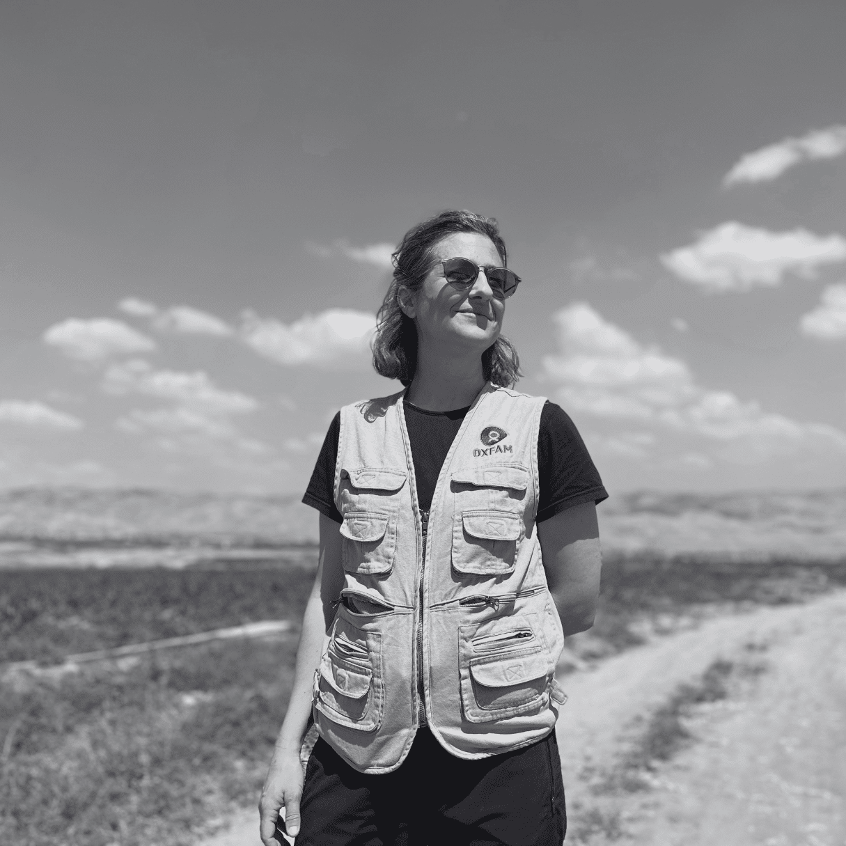 Lauren Ravon standing outdoors on a farm, wearing a beige utility vest over a dark shirt and sunglasses, with green fields, a dirt road, and distant hills under a blue sky in the background
