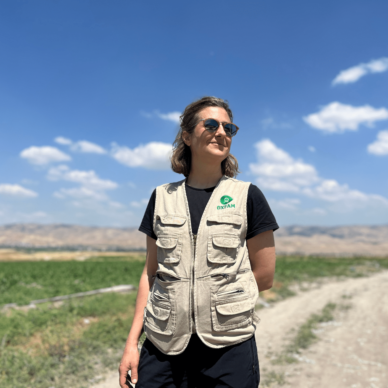 Lauren Ravon standing outdoors on a farm, wearing a beige utility vest over a dark shirt and sunglasses, with green fields, a dirt road, and distant hills under a blue sky in the background