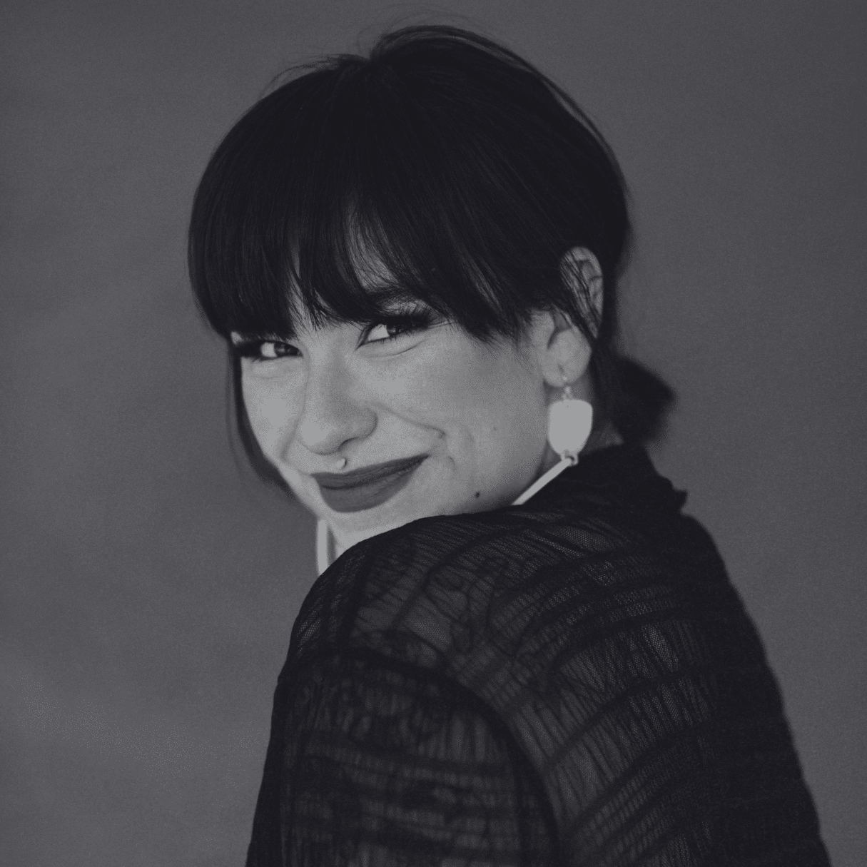 Portrait of Riley Yesno wearing a sheer black top and light colored earrings, turned slightly to the side against a solid red background