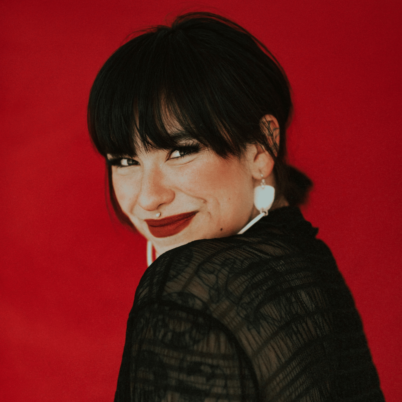Portrait of Riley Yesno wearing a sheer black top and light colored earrings, turned slightly to the side against a solid red background