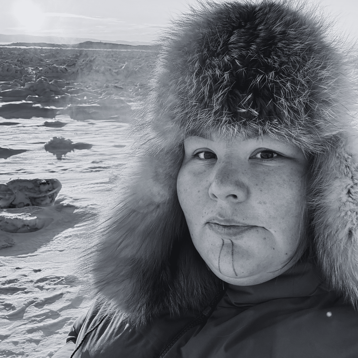 Rebecca (Becky) Mearns outdoors in a snowy landscape, wearing a thick winter coat and a fur trimmed hat, with ice formations and a low sun in the background