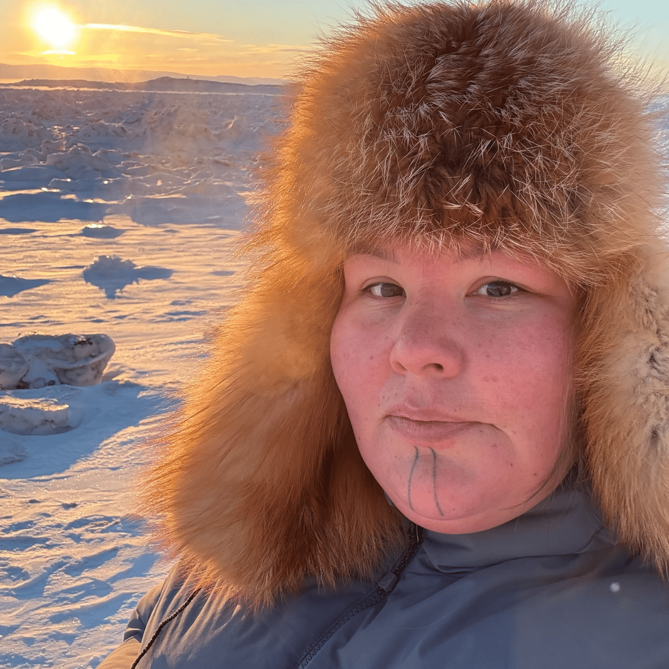 Rebecca (Becky) Mearns outdoors in a snowy landscape, wearing a thick winter coat and a fur trimmed hat, with ice formations and a low sun in the background