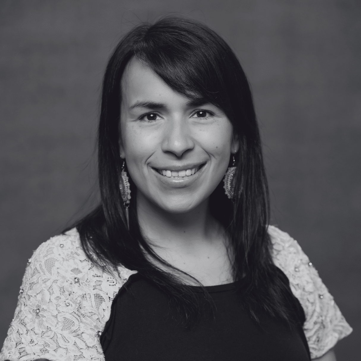 Studio portrait of Jayla Rousseau-Thomas wearing a black top with light colored lace sleeves and dangling earrings, standing against a neutral textured background