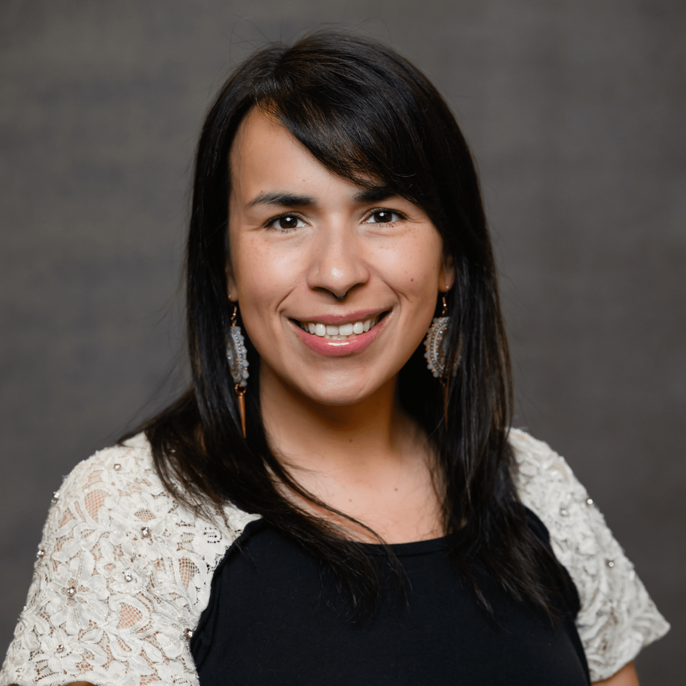 Studio portrait of Jayla Rousseau-Thomas wearing a black top with light colored lace sleeves and dangling earrings, standing against a neutral textured background