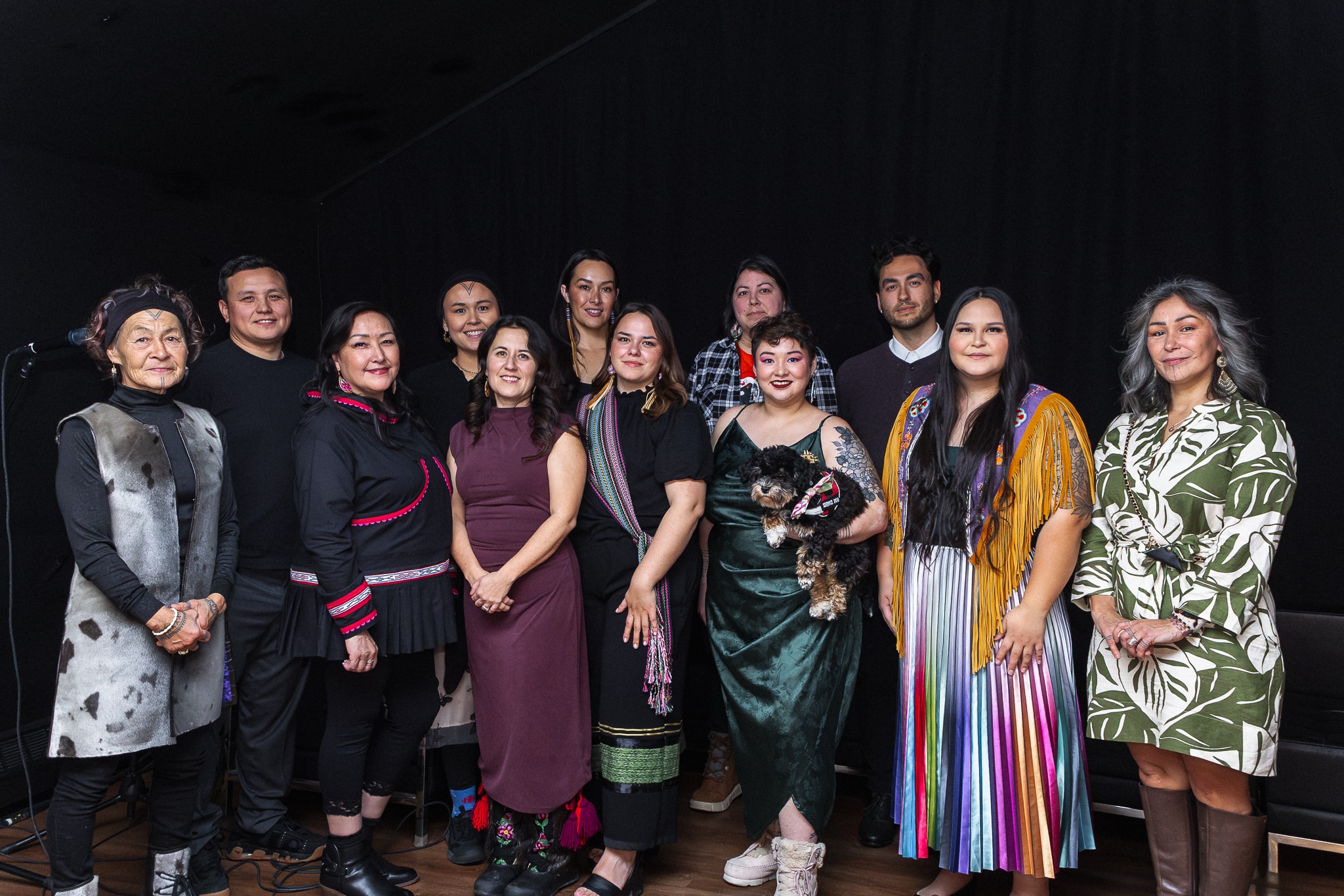 A group of twelve Indigenous entrepreneurs stand in a line smiling at the camera. One woman in front holds a small dog in her arms.