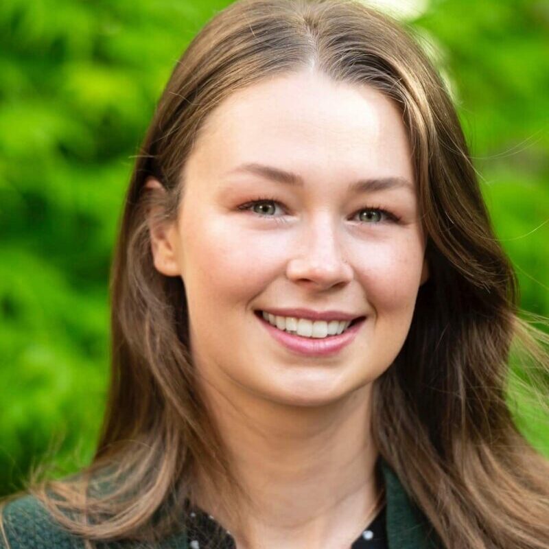 Chantelle posed in front of green nature background. She has light brown hair, and is wearing a forest green collared top.
