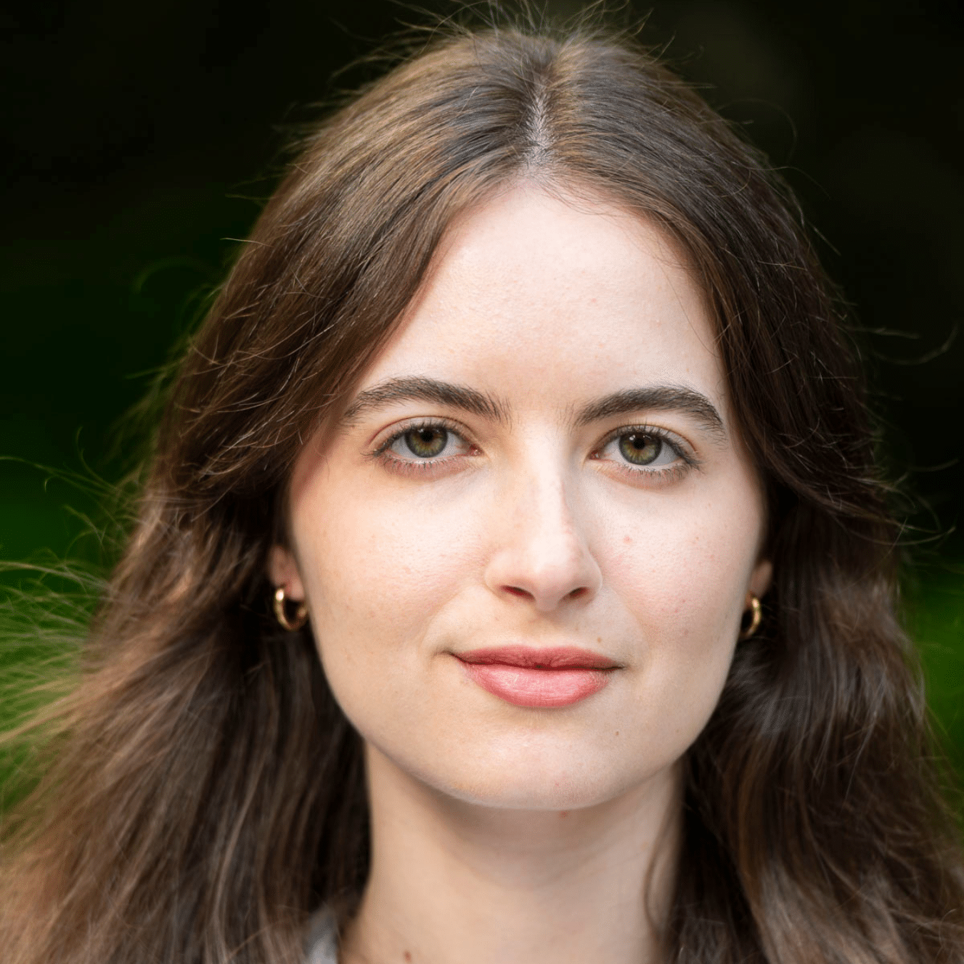 Headshot of Helena with long brown hair wearing a light blue and white striped shirt, standing outdoors against a softly blurred green background.