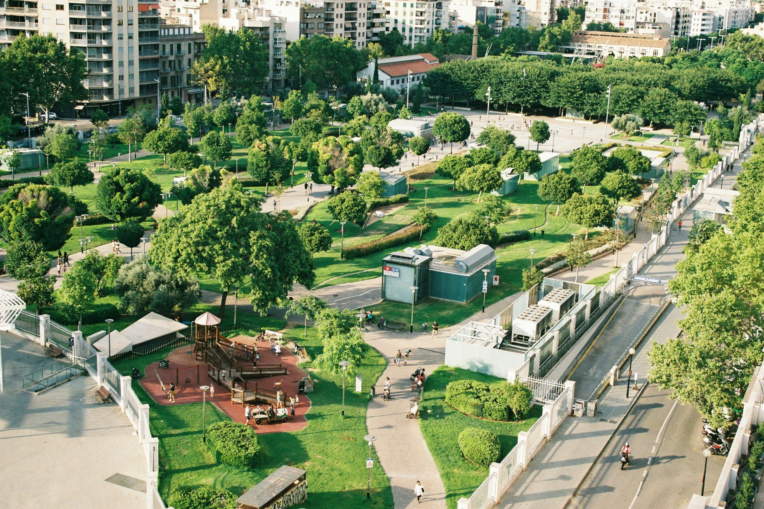 Aerial view of a large urban park with green lawns, tree-lined paths, a children's playground, and surrounding apartment buildings.