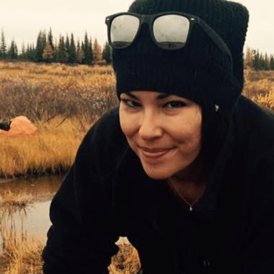 Ashley wearing a black beanie and dark clothing crouches beside a partially butchered moose carcass, in a grassy marsh area with trees in the background.