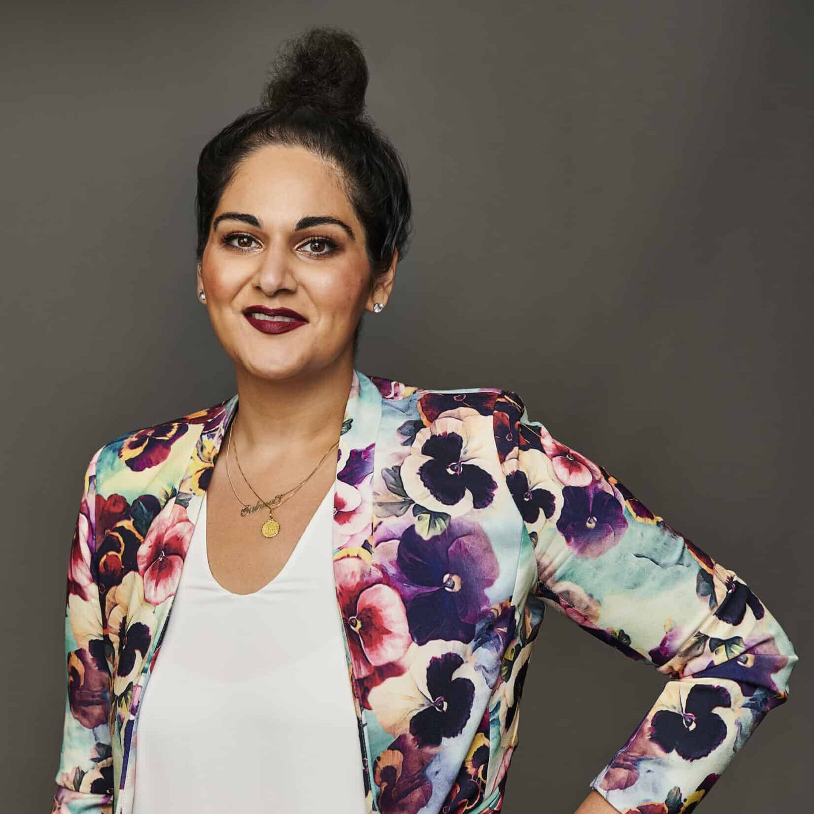 Professional headshot of Salma Dhanani standing against a dark grey background, wearing a colourful floral blazer over a white top, with hair styled in a high bun