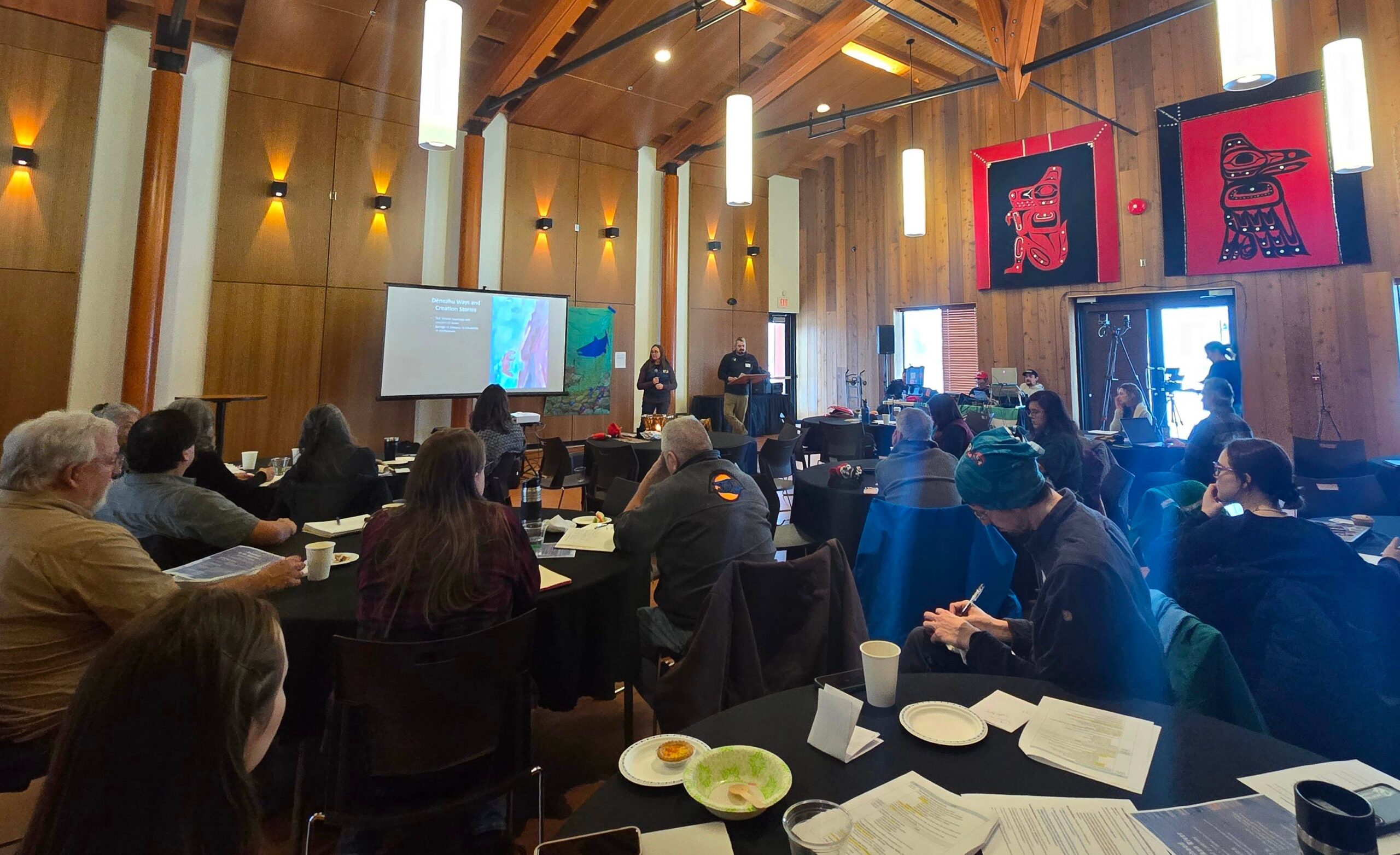 To Swim and Speak with Salmon conference participants seated at round tables in a large wood-paneled hall while two presenters speak beside a projection screen.