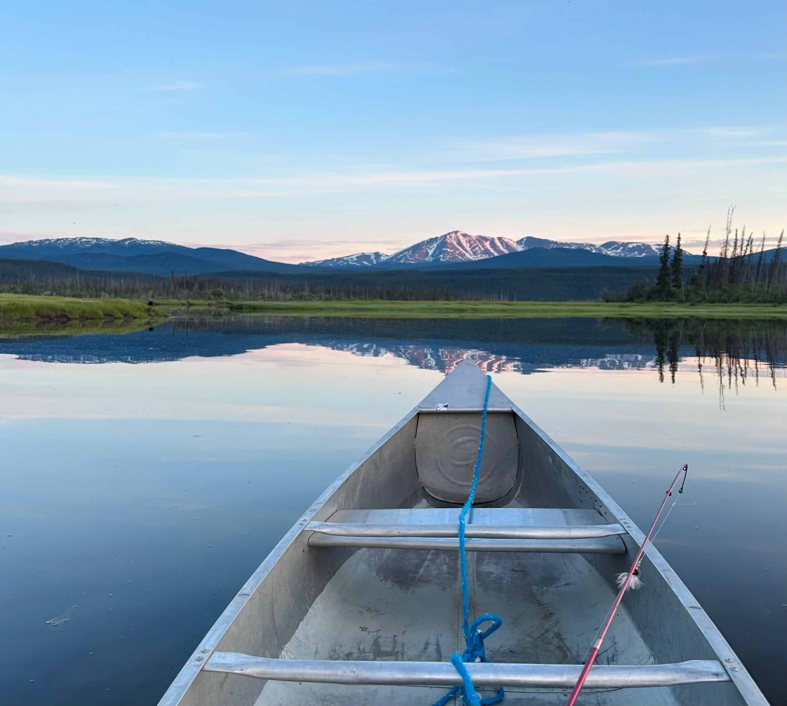 Canoe on a calm lake reflecting surrounding mountains and sky, with a snow-capped peak visible in the distance.